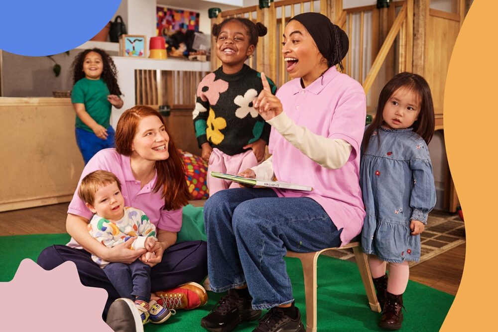 A woman sits on a chair ready to read a story surrounded by a group of children while another woman hugs a child.