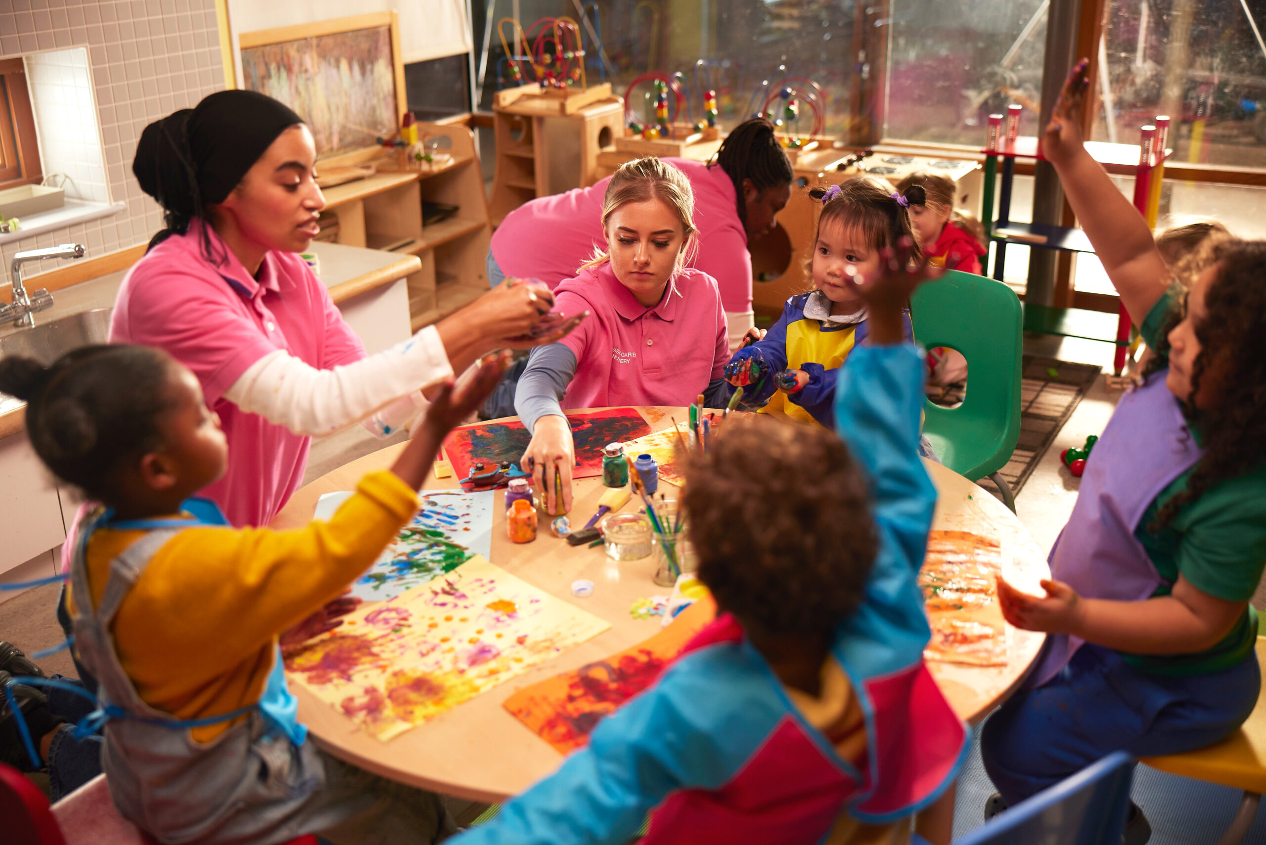 A group of children sitting with two women nursery staff around an arts and crafts table in a playroom.